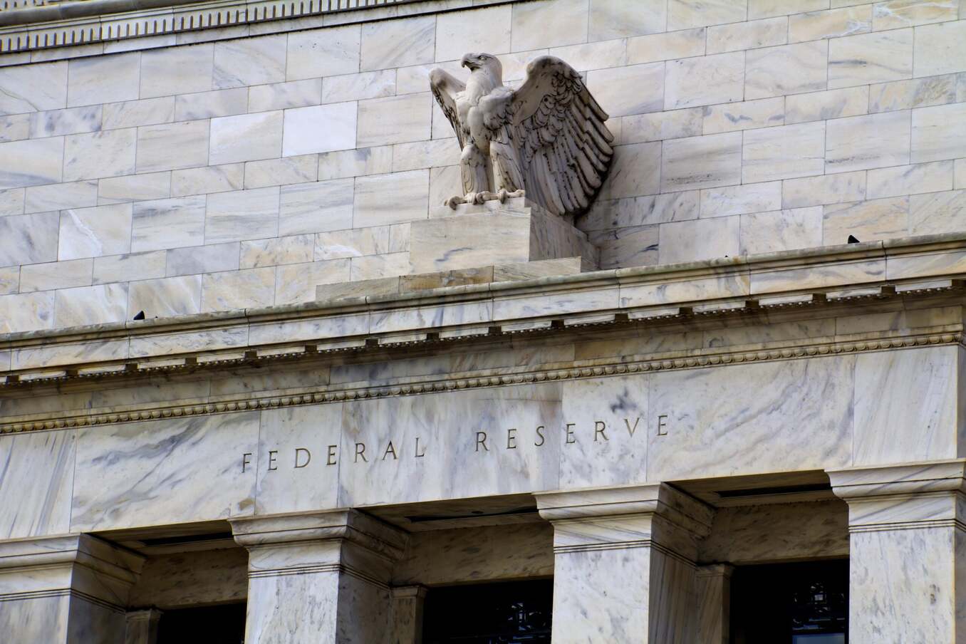 Exterior of the Federal Reserve building showing the word "FEDERAL RESERVE" carved into the marble facade beneath a stone eagle statue