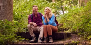 A smiling middle-aged man and woman taking a break during a hike, sitting together on rustic wooden stairs in a lush, sunlit green forest. The man wears a red plaid shirt and backpack, while the woman wears a blue long-sleeve shirt and looks at him admiringly. Both are wearing sturdy hiking boots.