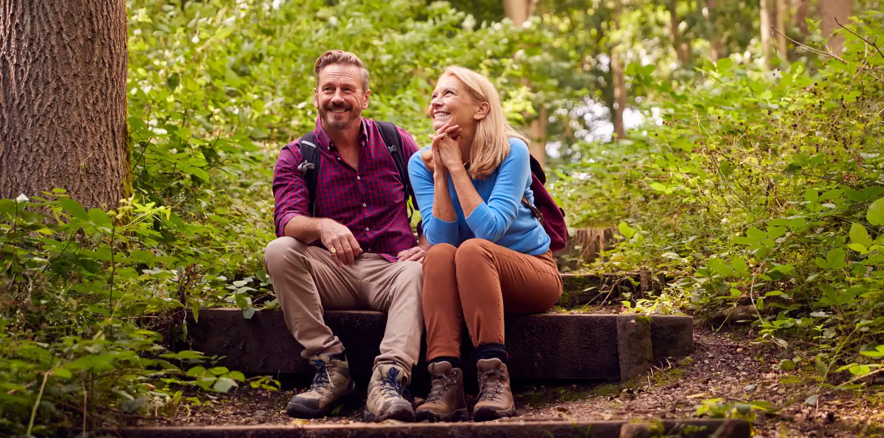 A smiling middle-aged man and woman taking a break during a hike, sitting together on rustic wooden stairs in a lush, sunlit green forest. The man wears a red plaid shirt and backpack, while the woman wears a blue long-sleeve shirt and looks at him admiringly. Both are wearing sturdy hiking boots.
