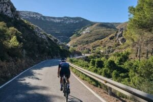 A cyclist in professional gear rides along a winding asphalt road through a lush mountain valley. The scene features steep, rocky hillsides dotted with Mediterranean vegetation and a clear blue sky.