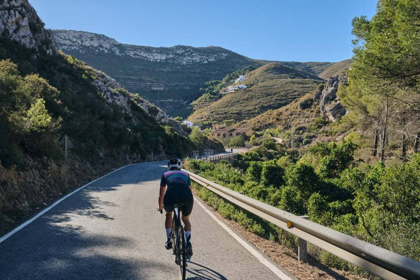A cyclist in professional gear rides along a winding asphalt road through a lush mountain valley. The scene features steep, rocky hillsides dotted with Mediterranean vegetation and a clear blue sky.