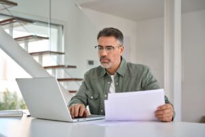 A middle-aged man with glasses sitting at a table, focused on his laptop while holding several sheets of paper in his home office