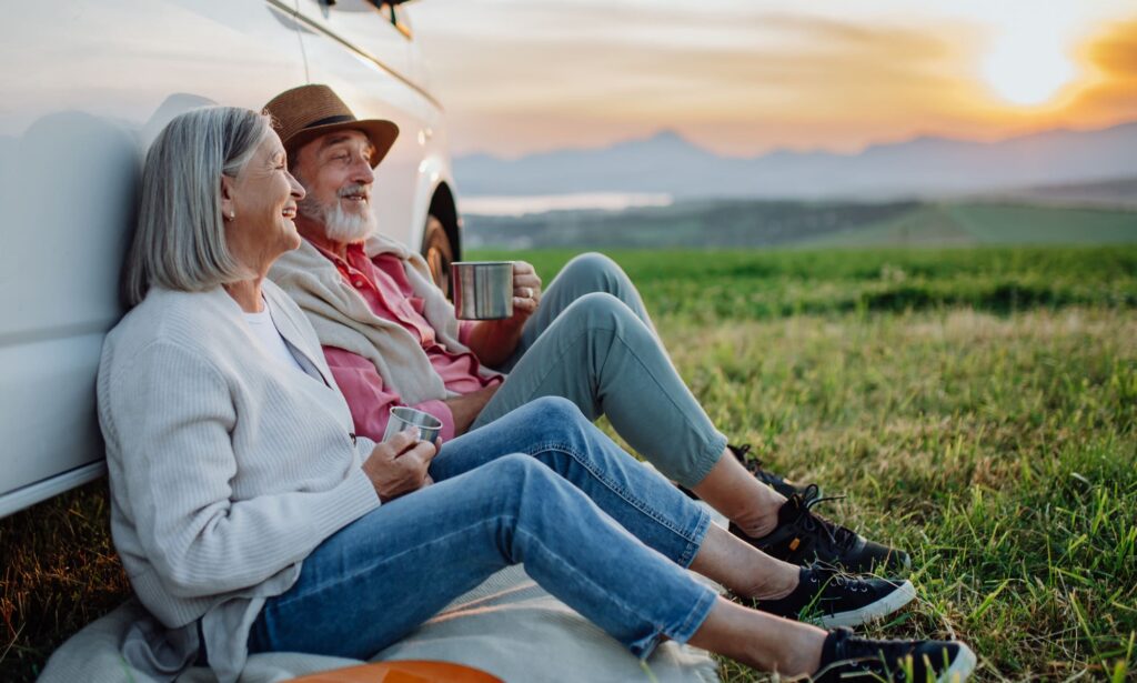 Potrait of senior couple sitting by car and drinking coffee after long drive during their roadtrip. Elderly spouses at autumn roadtrip watching the sun set behind High Tatras