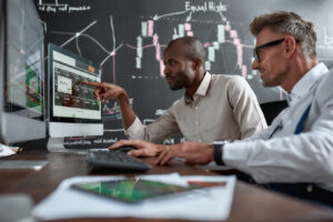Two diverse colleagues traders talking to each other, looking at graphs while sitting in the office in front of multiple computer screens.