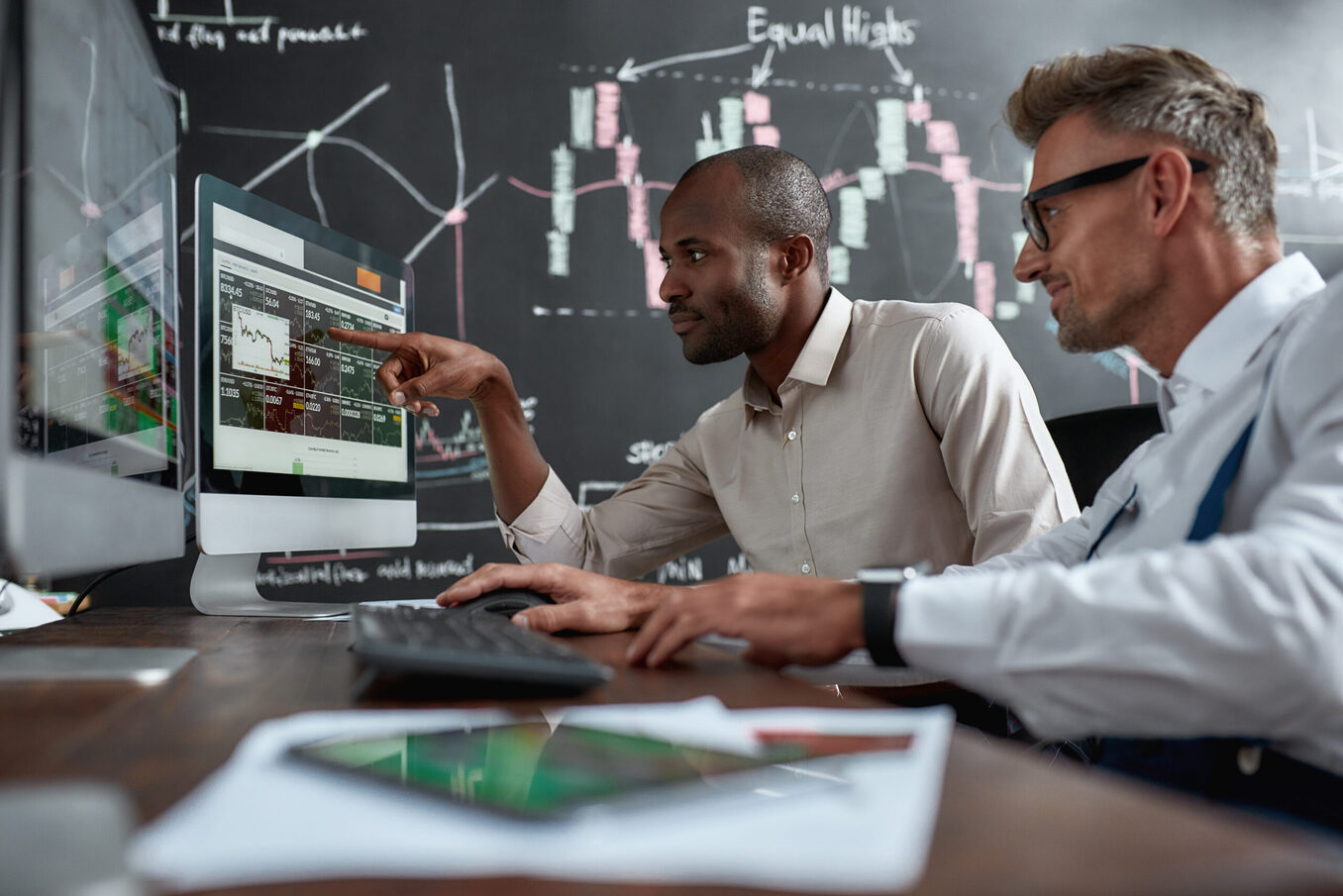 Two diverse colleagues traders talking to each other, looking at graphs while sitting in the office in front of multiple computer screens.