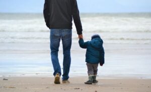Father holding hands with his young child while walking along a sandy beach toward the ocean waves on a cool day.
