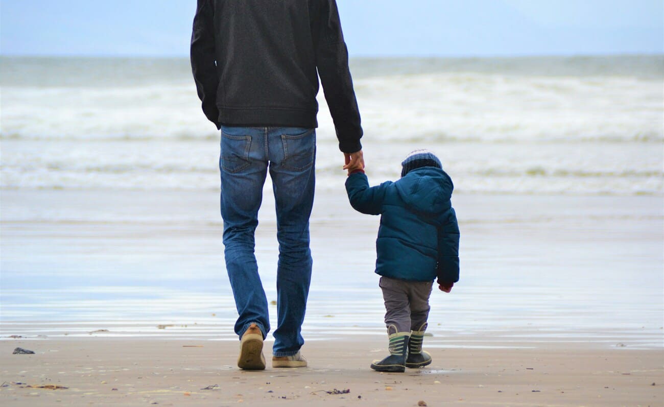 Father holding hands with his young child while walking along a sandy beach toward the ocean waves on a cool day.
