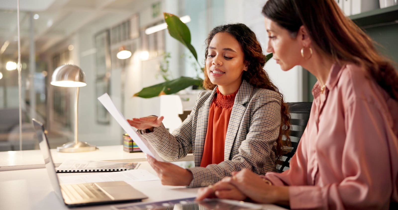 Two female professionals reviewing business strategy report and laptop in modern office