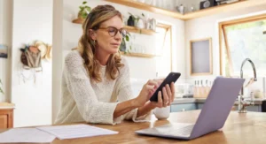 A mature woman wearing glasses sitting in a brightly lit kitchen, using her smartphone while working on a laptop with financial documents nearby to review Medicare IRMAA costs.
