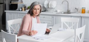 A senior woman sitting at a kitchen table reviewing financial paperwork and investment documents while using a laptop.