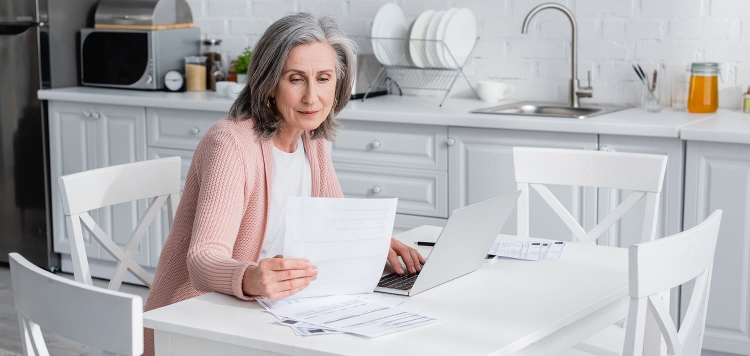 A senior woman sitting at a kitchen table reviewing financial paperwork and investment documents while using a laptop.