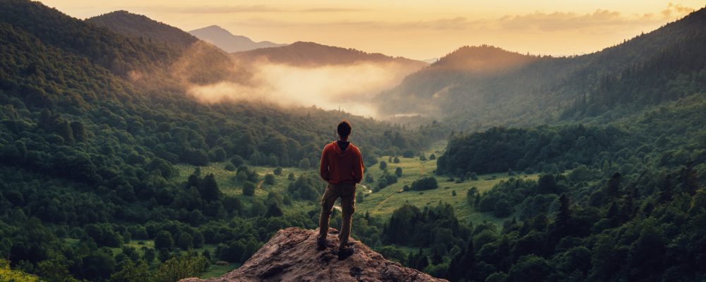 Young man standing on top of cliff in summer mountains at sunset and enjoying view of nature