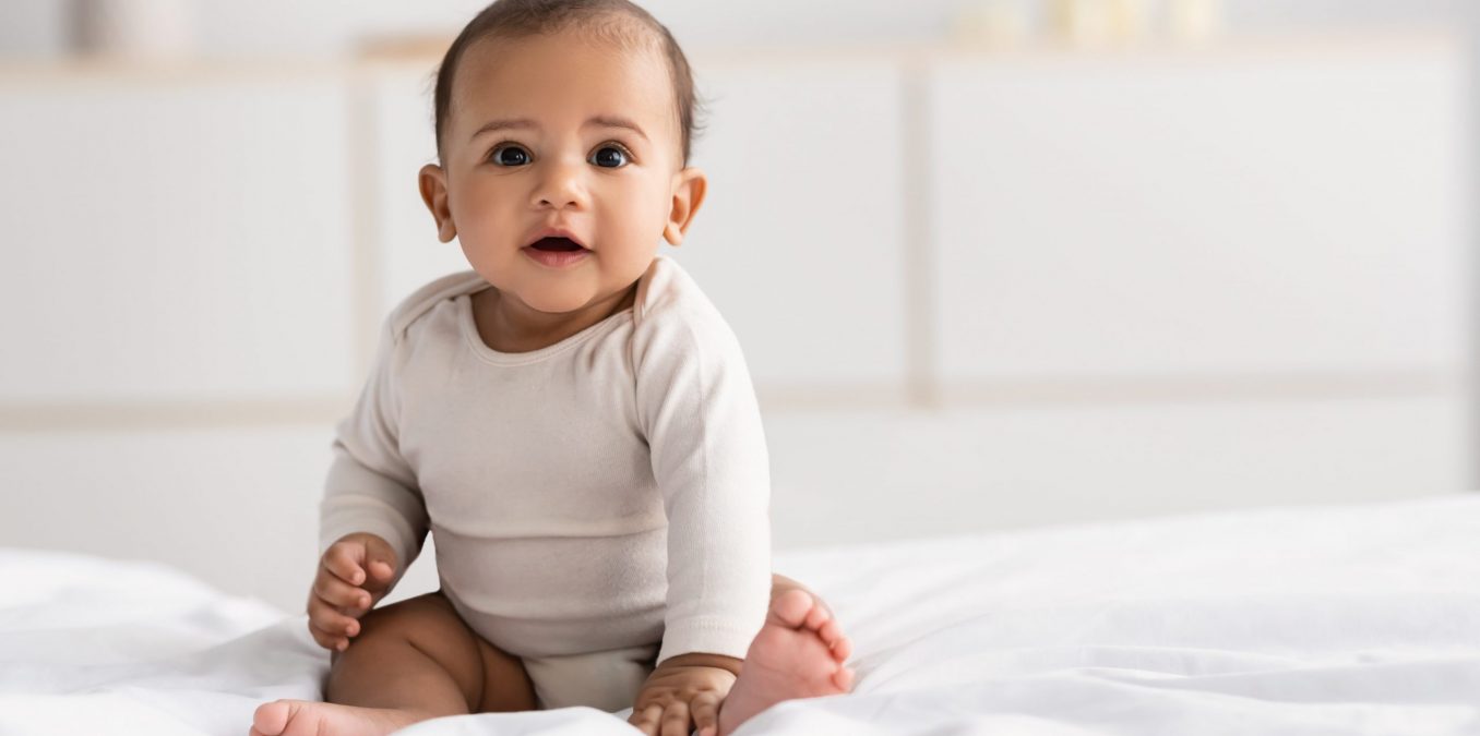 An adorable baby sitting on a white bed in a bright, modern room.