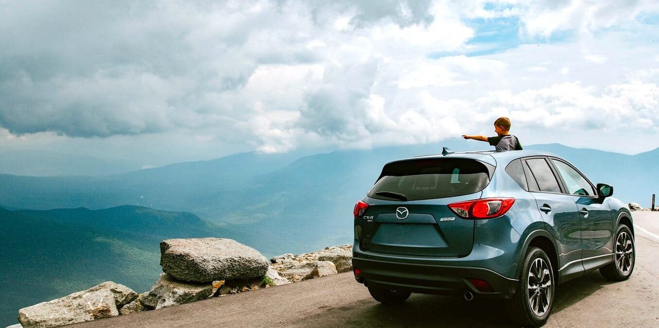 A young boy looking out from the sunroof of a blue SUV parked at a mountain overlook, pointing toward a vast valley under a cloudy sky