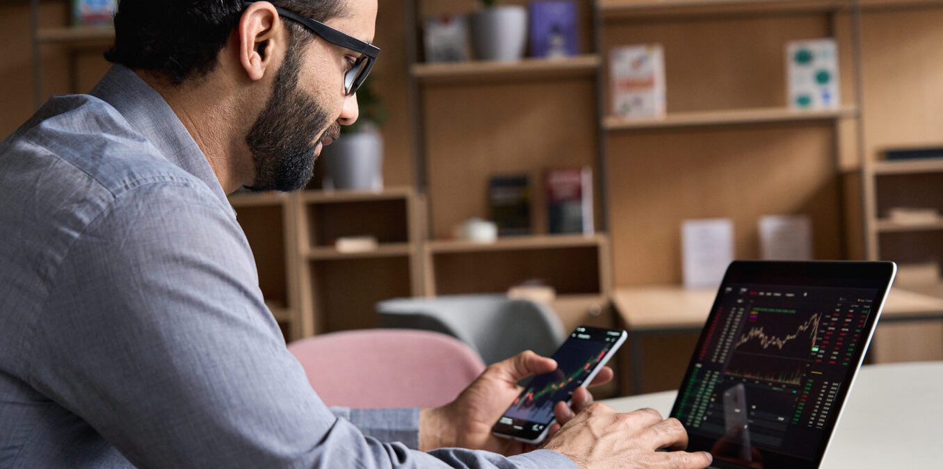Indian latin business man trader investor analyst looking at financial stock market charts rate dynamic, trading data indexes growth using mobile phone and laptop for cryptocurrency digital analytics.