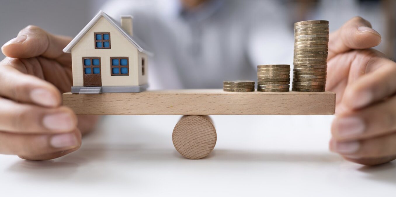 Businessperson's Hand Protecting Balance Between House Model And Increasing Stacked Coins On Seesaw