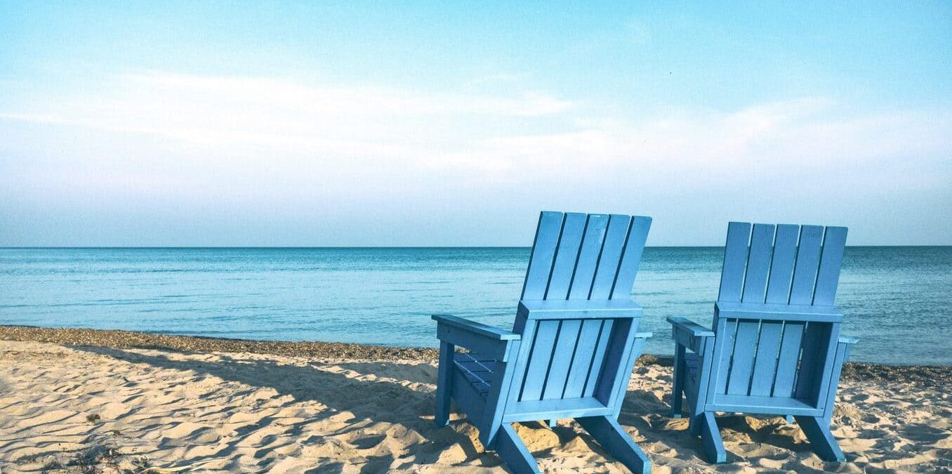 Two empty light blue Adirondack chairs sitting on a sandy beach, facing a calm, turquoise ocean under a clear blue sky. The scene is peaceful and sunny, with soft shadows on the sand.