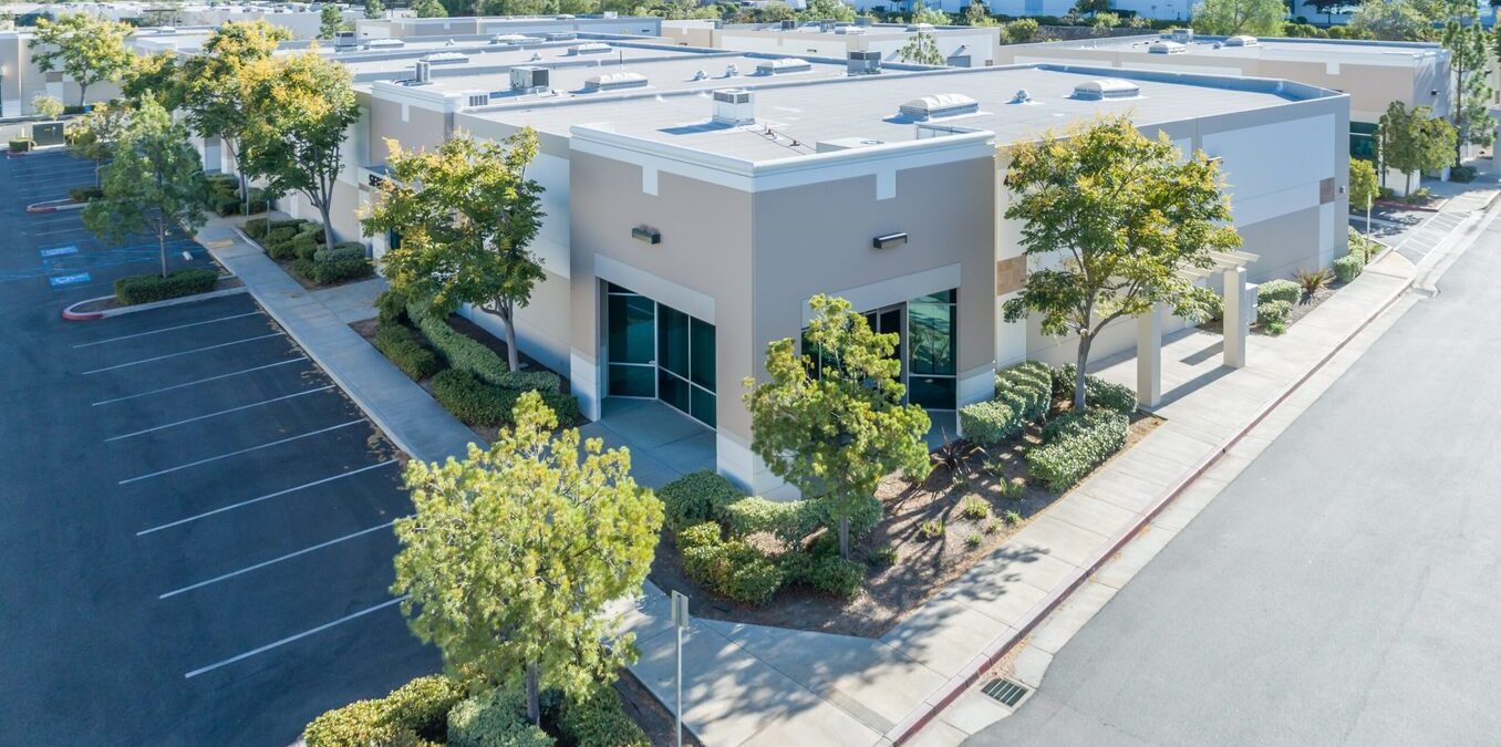 An aerial high-angle view of a modern one-story commercial office building in a suburban business park. The building features beige and white exterior walls with large glass windows, surrounded by paved parking lots, green trees, and manicured landscaping under a clear sky.