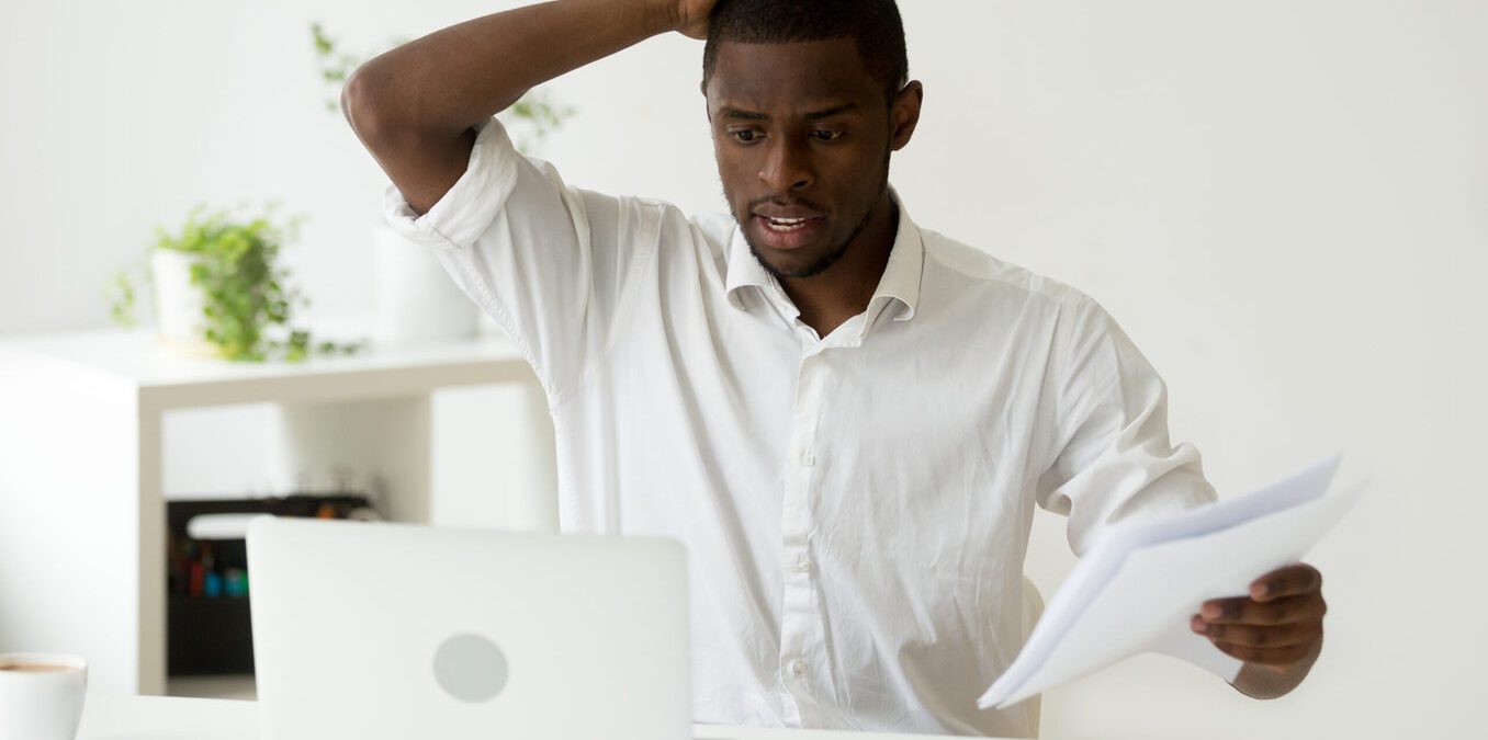 Confused African-American businessman having problem with documents looking at laptop