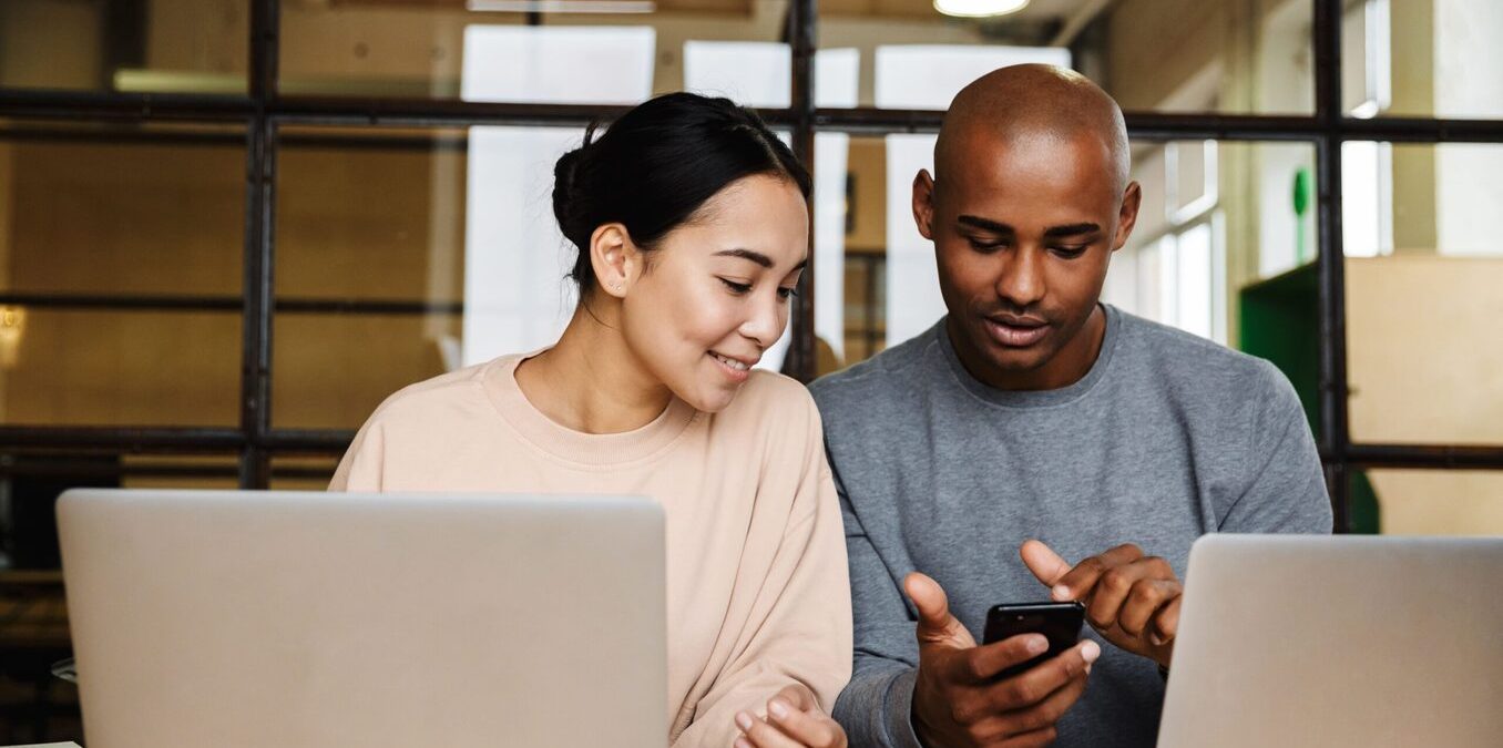 Couple in front of their laptops on a table. The man is holding a phone and showing something to his partner.