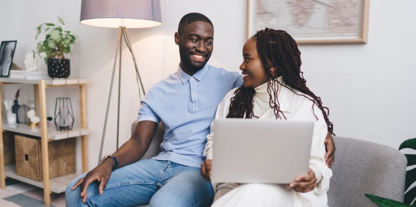 A smiling young couple sitting on a sofa and using a laptop together at home.