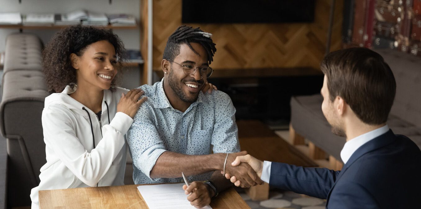 A smiling couple sitting at a wooden table, with the man shaking hands with a professional advisor across from them after signing a document