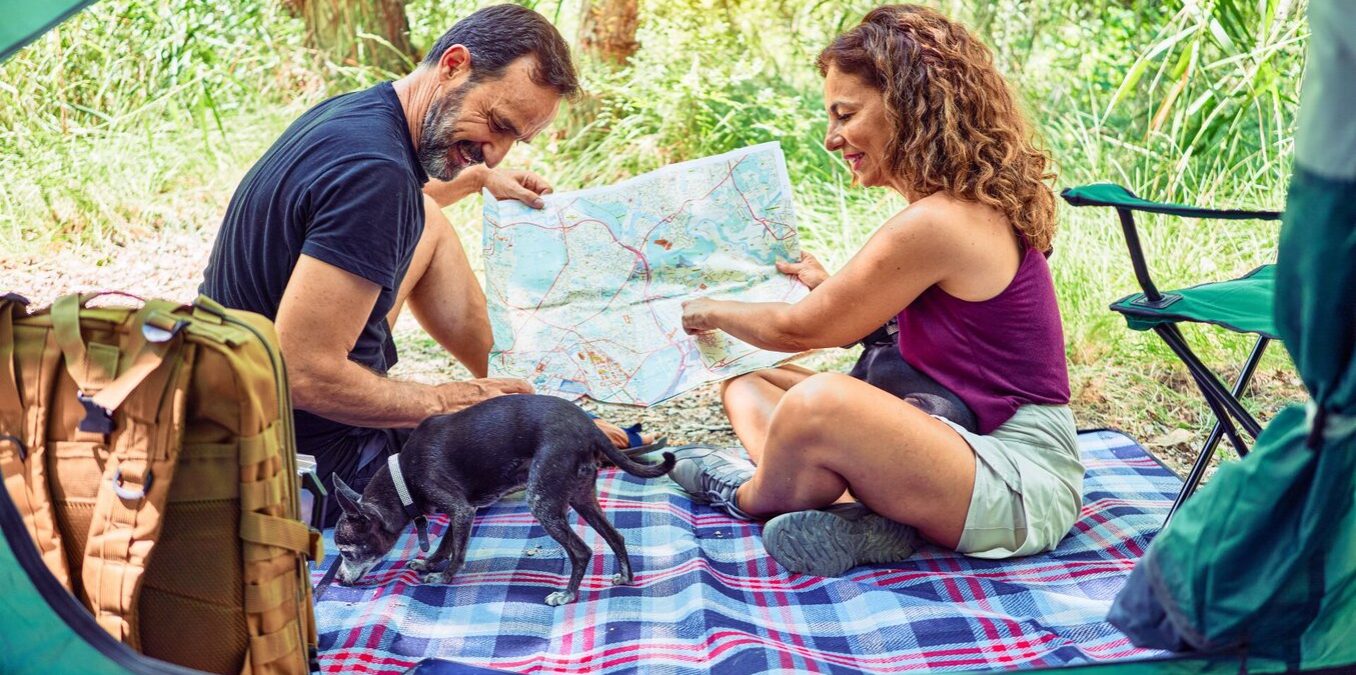 A middle-aged man and woman sitting on a blue plaid blanket outside their tent while camping in a forest. They are smiling and looking at a large paper map together, while a small black dog explores the blanket near their hiking gear and backpacks.