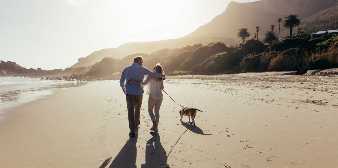 A view from behind of a middle-aged couple walking arm-in-arm along a wide, sandy beach at sunset. They are walking a beagle on a leash toward a backdrop of lush green hills and palm trees, with the warm sun creating long shadows on the sand.