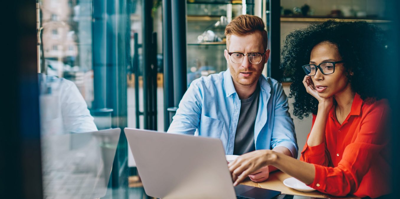 A diverse man and woman sitting at a small wooden table in a cafe, looking intently at a laptop screen together. The man has red hair and glasses, wearing a light blue shirt, while the woman has dark curly hair and glasses, wearing a vibrant orange blouse and pointing at the screen.
