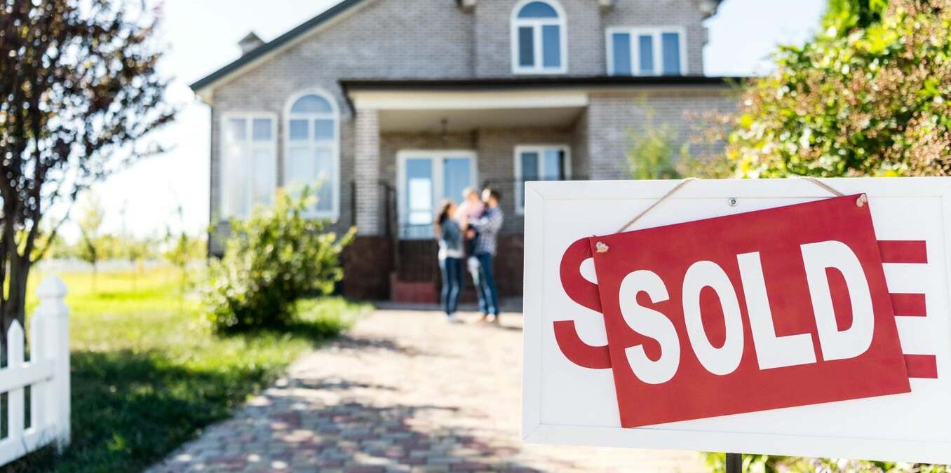 A close-up of a white real estate sign with a red "SOLD" placard in the foreground. In the blurred background, a happy family of three stands in front of a modern two-story brick house, celebrating their new home purchase on a sunny day.
