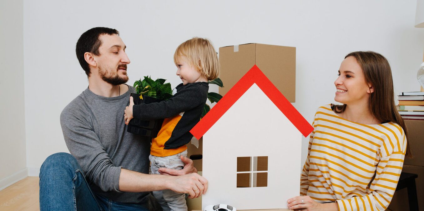 Family moved into a new apartment, parents looking at their son. They surrounded by multiple sealed cardboard boxes. Mother holding toy paper house in hands, symbol of a new home.