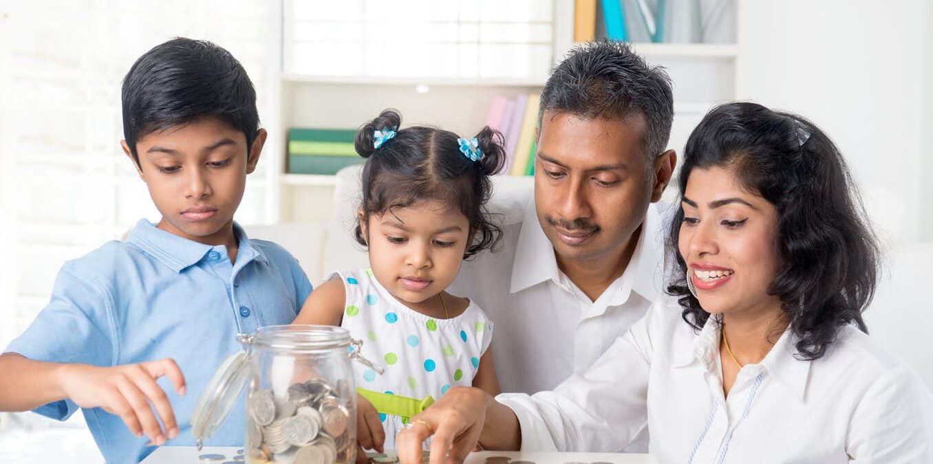 A family of four sitting together at a table, with two young children and their parents counting coins and putting them into a glass savings jar