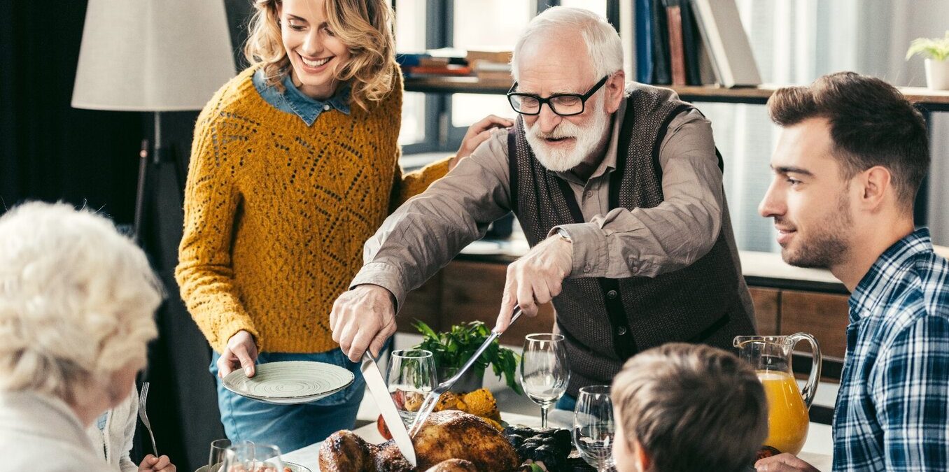 A senior man with glasses and a white beard carving a roasted turkey at a crowded dinner table. His family, including a young woman in a yellow sweater holding a plate and a man in a plaid shirt, watch with smiles. The scene is a warm, indoor family gathering with wine glasses and side dishes visible.