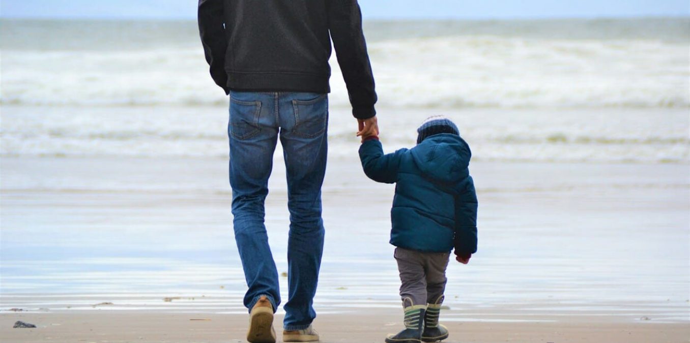 Father holding hands with his young child while walking along a sandy beach toward the ocean waves on a cool day.