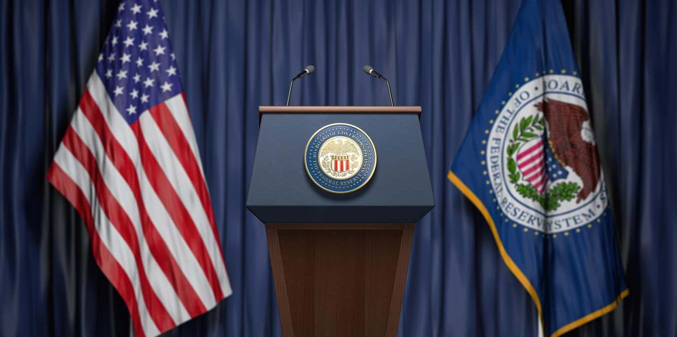 A wooden podium featuring the Federal Reserve seal, flanked by the United States flag and the Federal Reserve System flag against a dark blue curtain backdrop