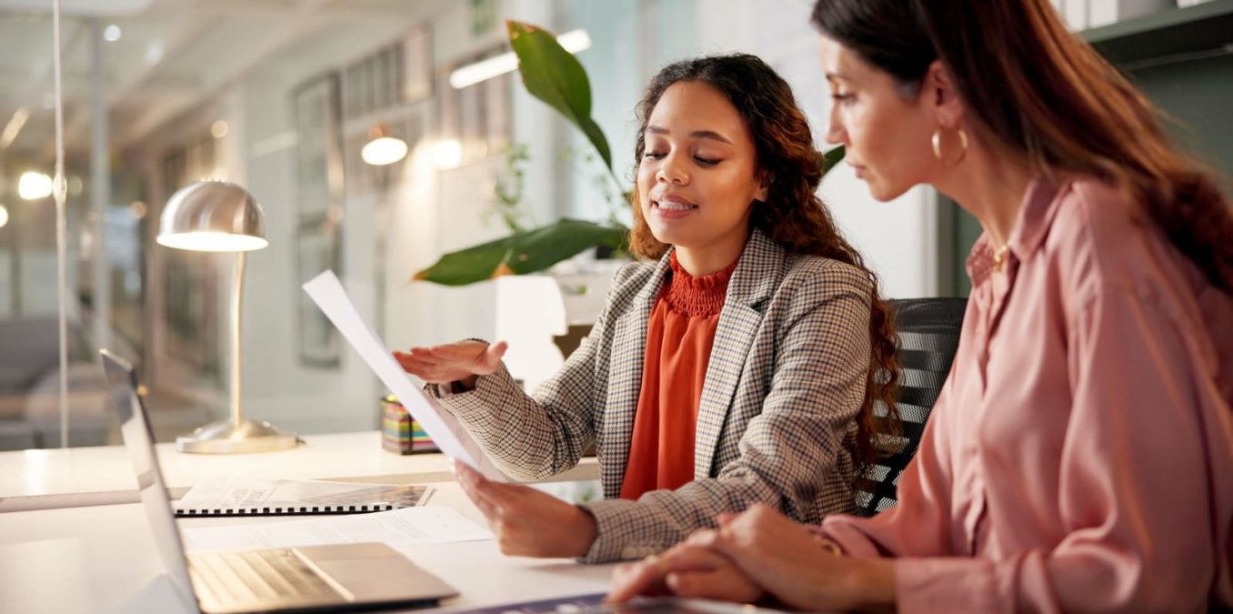 Two female professionals reviewing business strategy report and laptop in modern office