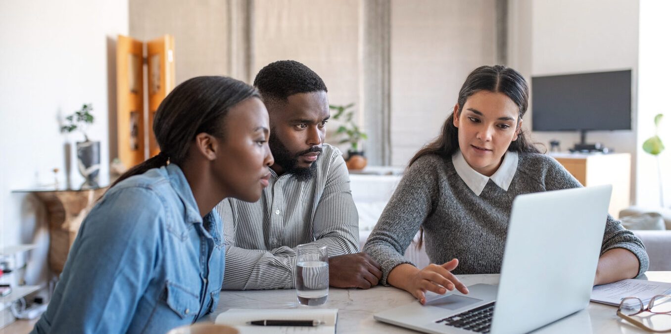 Financial planner going over savings plans on a laptop with a young African American couple at a table in their living room at home