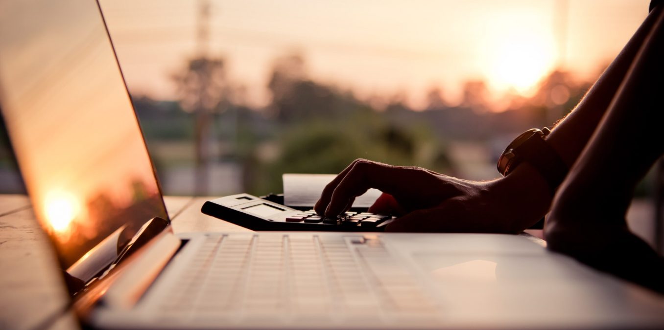 Close-up of a person using a laptop and calculator outdoors during a warm sunset.