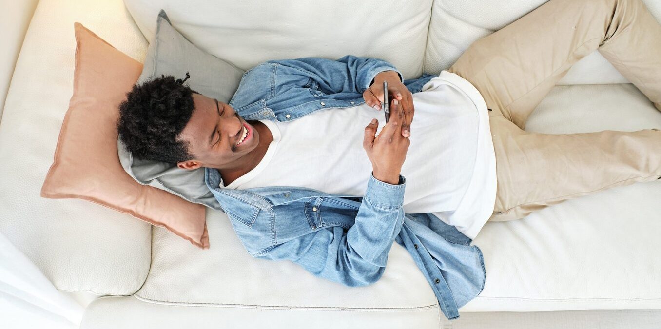 Man lying down on couch, looking at his phone and laughing.