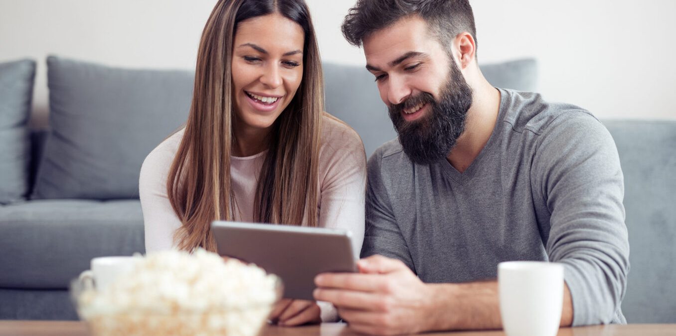 A happy couple sitting at home and watching content on a tablet with popcorn.