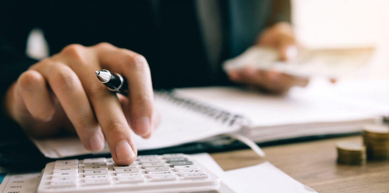 A close-up of a person's hands using a white calculator while holding a pen over an open notebook. In the blurred background, they are holding a fan of cash next to stacks of coins on a wooden desk, symbolizing accounting, budgeting, or financial management.