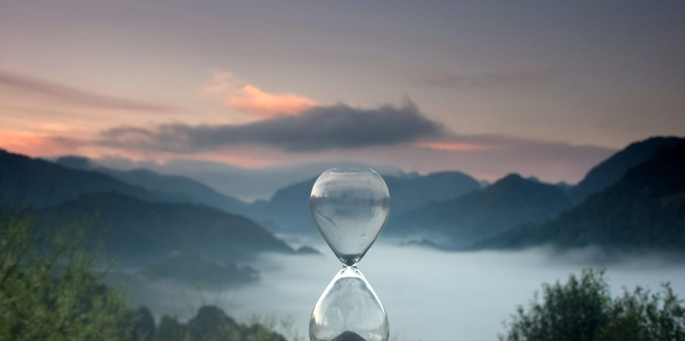 A hourglass (with falling sand) on a wooden wet table with sea of fog and mountain range silhouette in sunrise time
