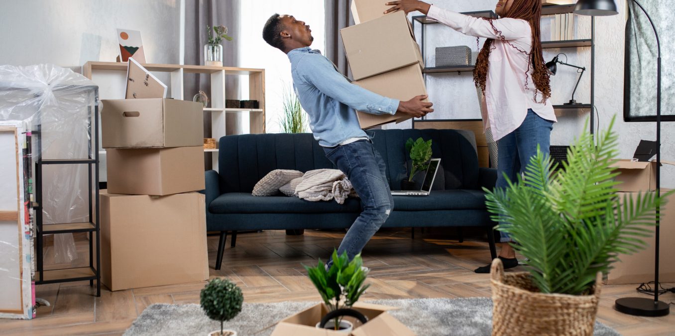 A young couple playfully struggling with a stack of heavy boxes while moving into a new home.