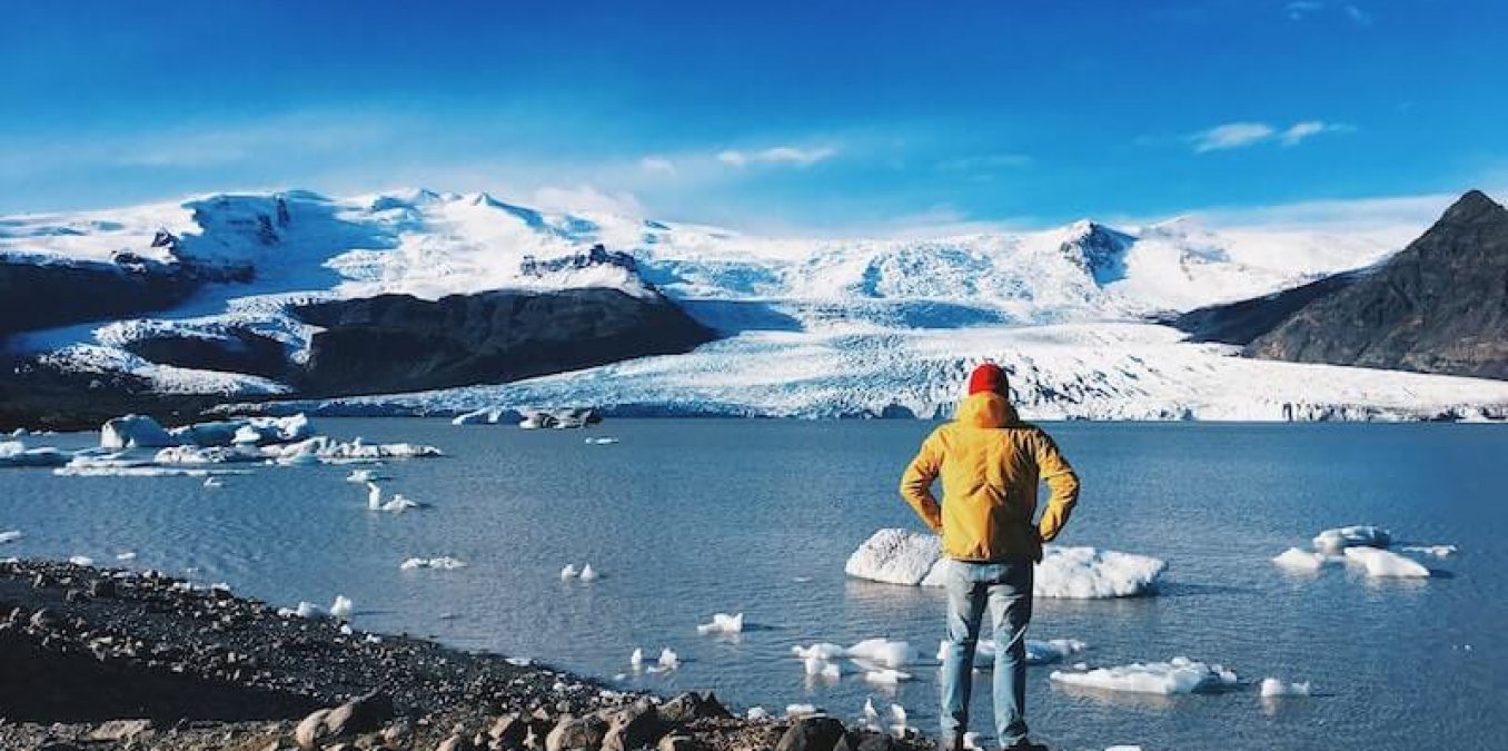 A person in a yellow jacket and red hat standing on a rocky shore, looking out at a glacial lake with icebergs and snow-covered mountains in the background