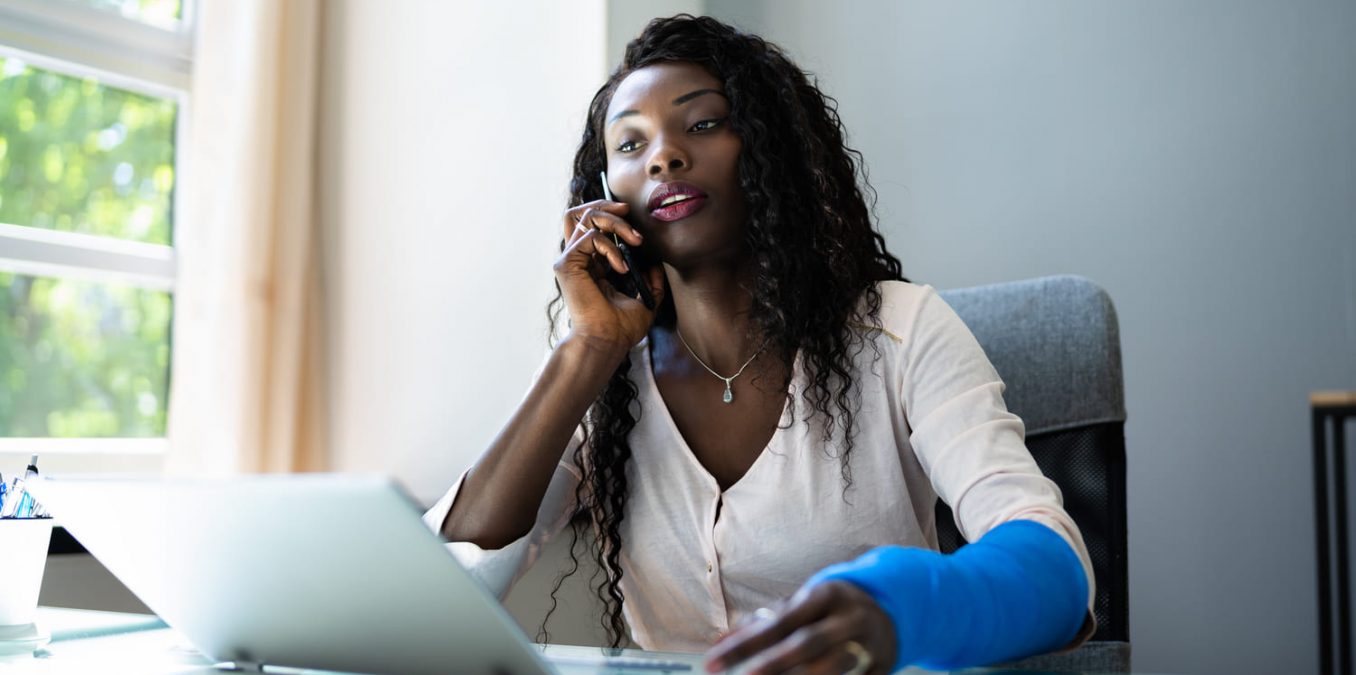 An office worker with her arm in a blue cast talking on the phone at her desk.