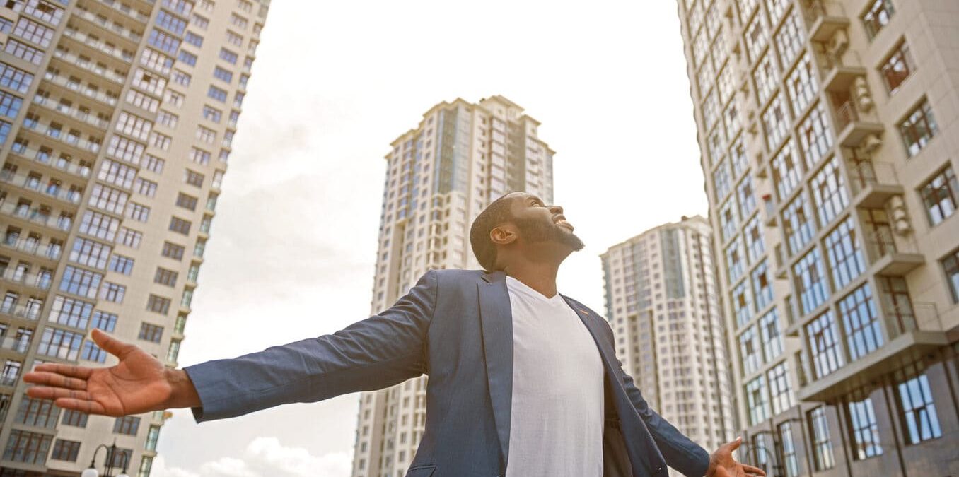 A man in a blue blazer standing with his arms outstretched and head tilted back, looking up toward tall apartment buildings under a bright sky