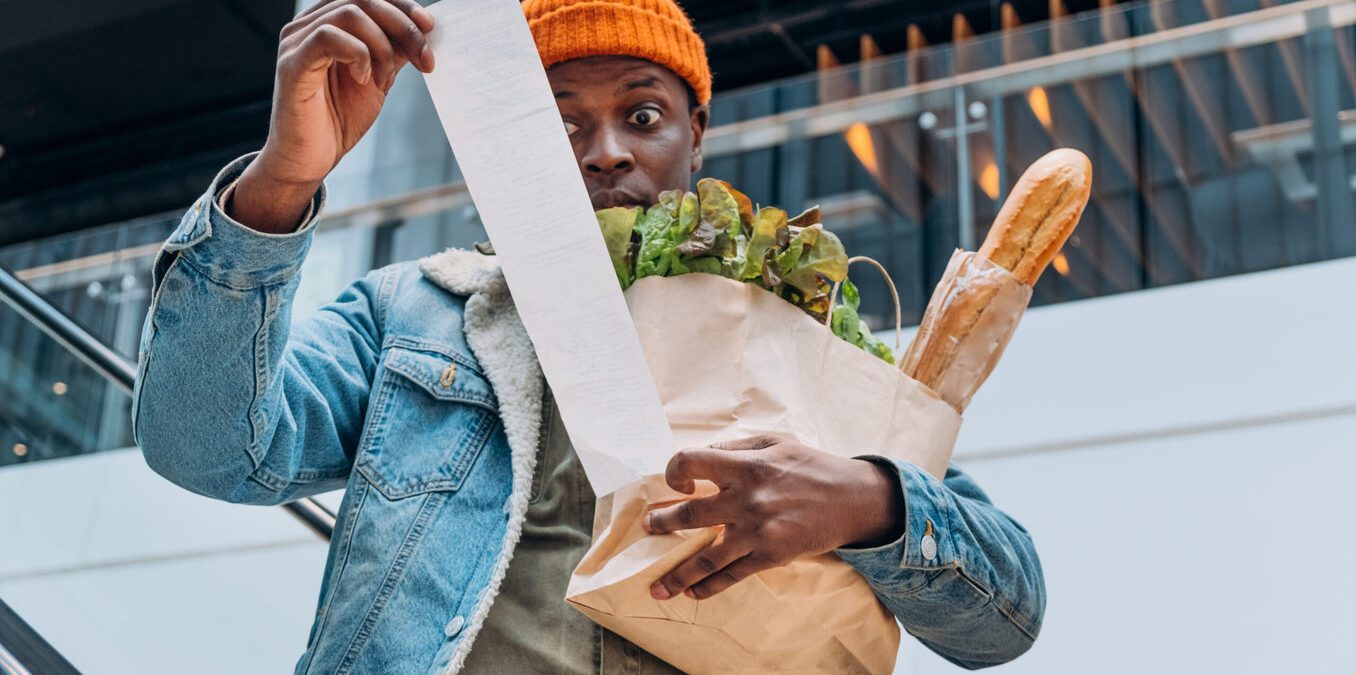 A man with a surprised expression holding a brown paper bag of groceries and looking at a very long receipt