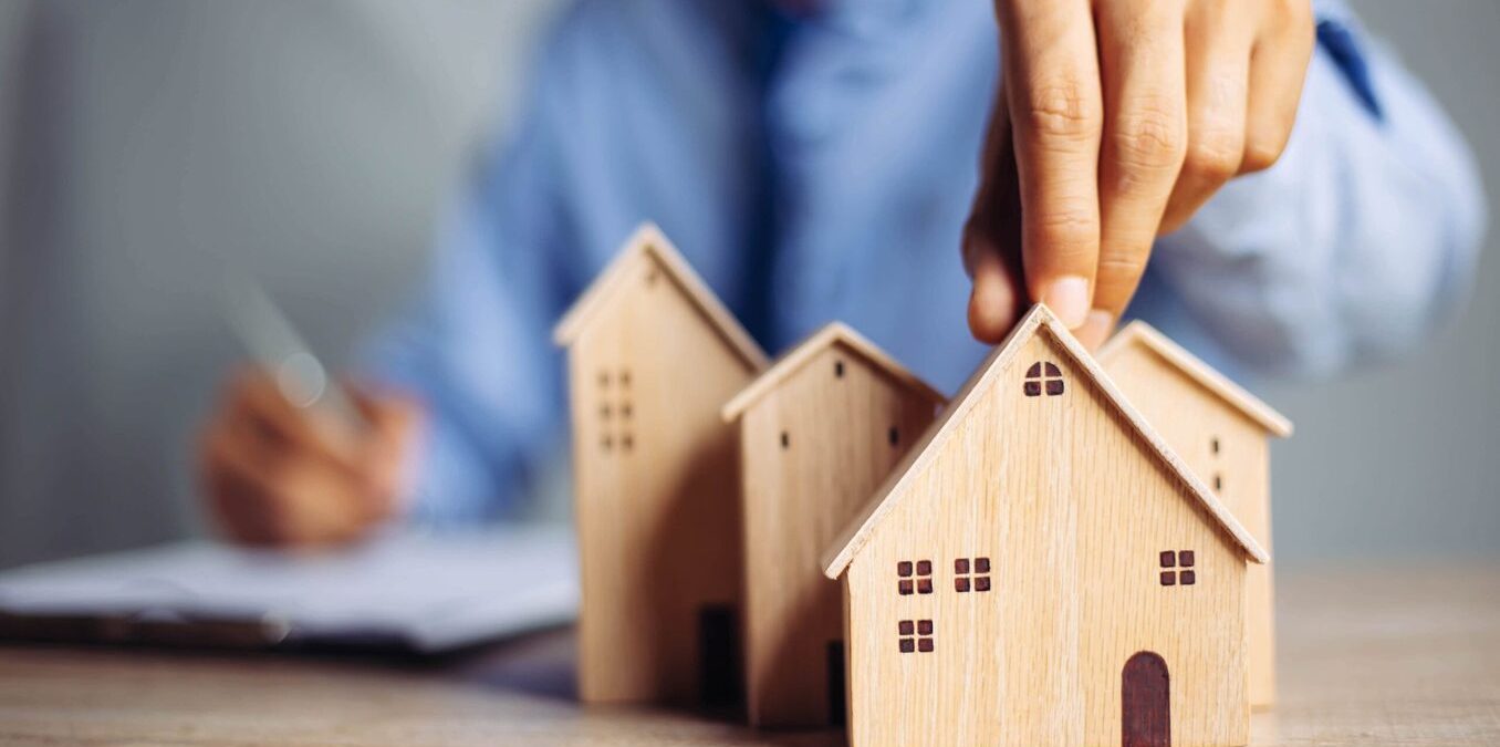A close-up of a person’s hand placing a small wooden house model among a cluster of other wooden houses on a desk. In the blurred background, a person in a blue shirt is writing on a clipboard, symbolizing real estate planning, home buying, or property investment.