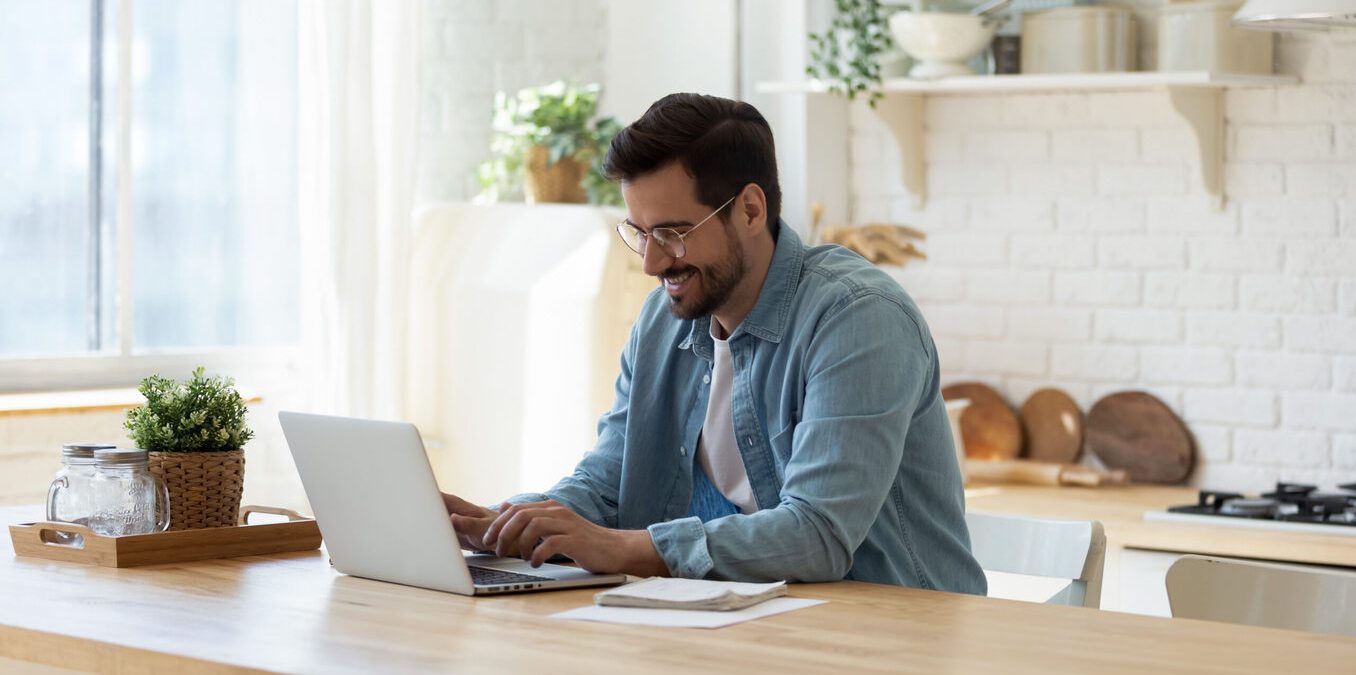 Focused millennial Caucasian man sit at table at home kitchen look at laptop screen working distant. Young male use computer, texting or messaging online on device. Communication, technology concept.