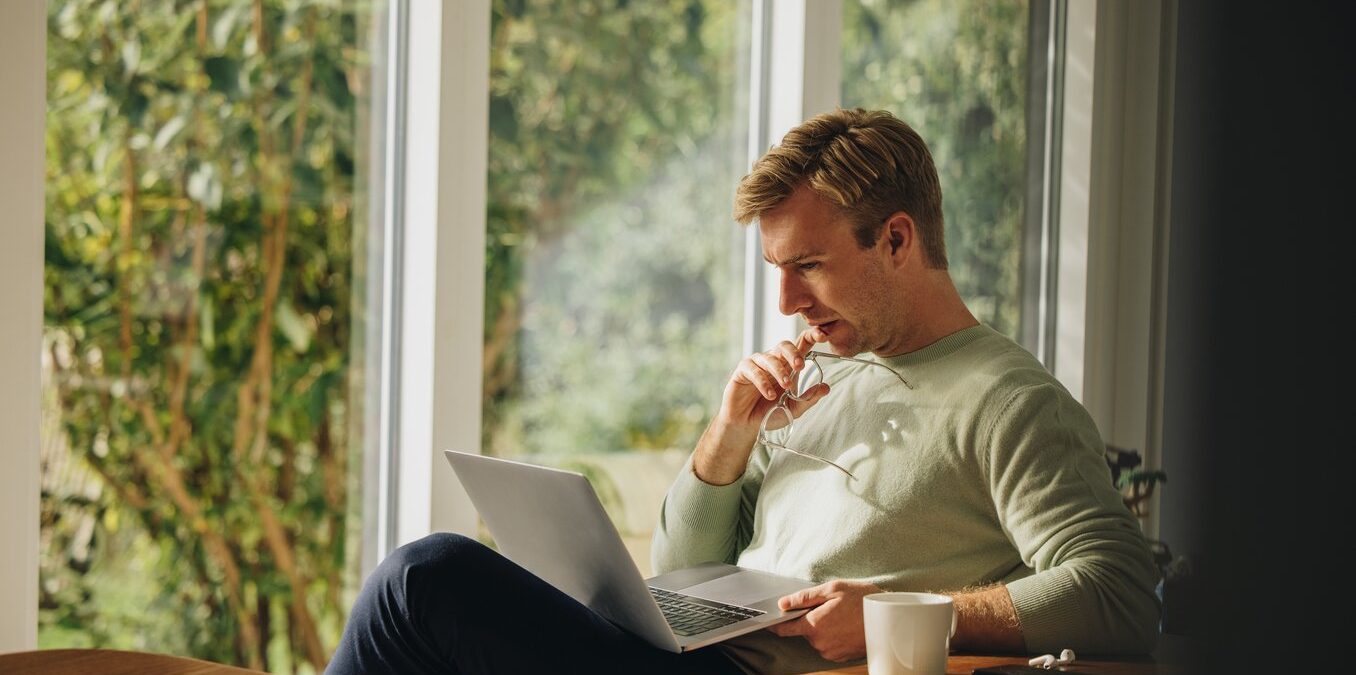 Man working on laptop and thinking. Man sitting at home looking at laptop and thinking.